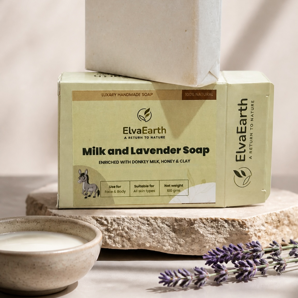 A bar of ElvaEarth Milk and Lavender Soap stacked on its branded cream-colored packaging box, styled on a stone slab with a small bowl of soap and lavender sprigs in a warm, sunlit setting.