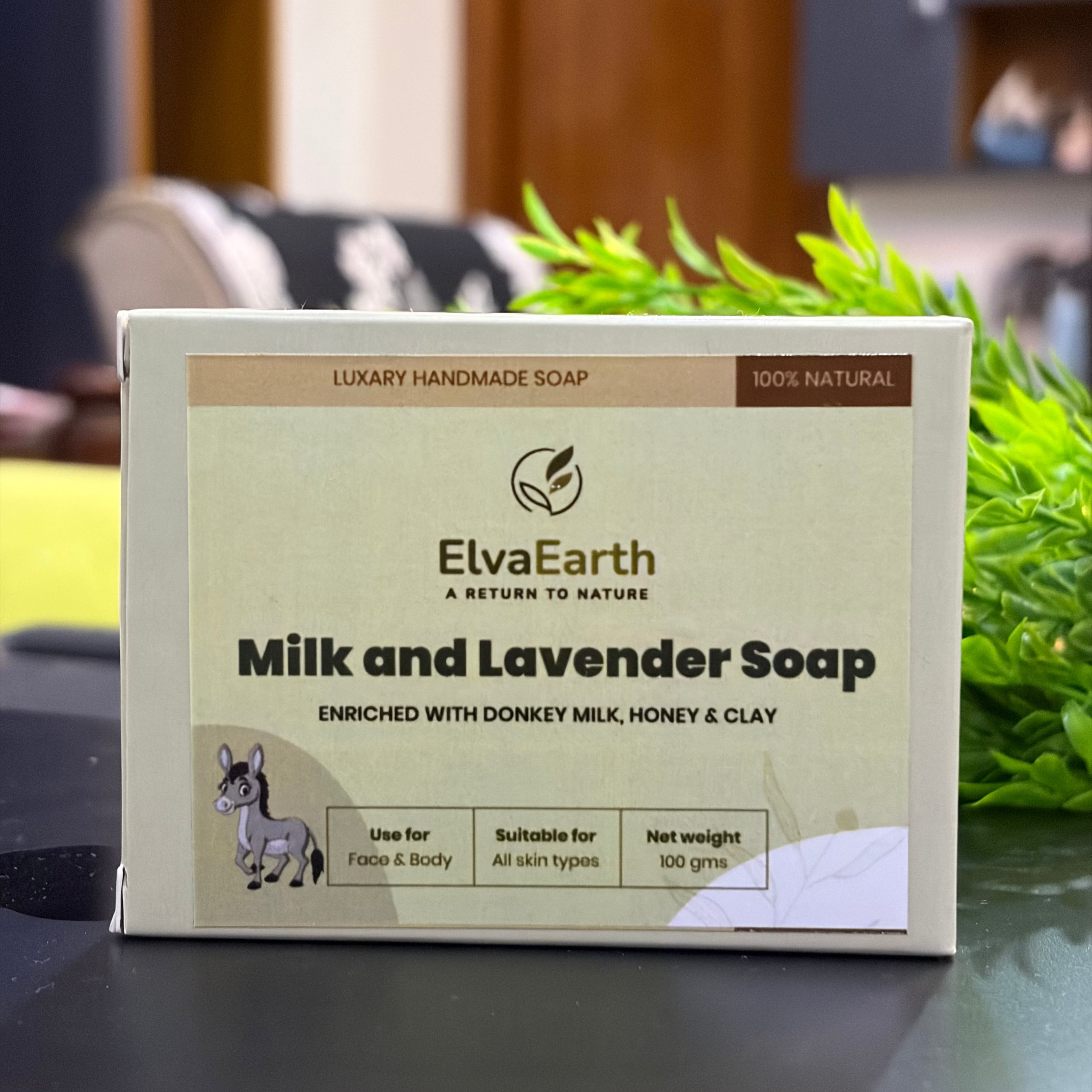 A rectangular box of ElvaEarth Milk and Lavender Soap sits on a dark tabletop with a blurred indoor background and green foliage behind it. The box is cream and tan with black text and a small donkey illustration.