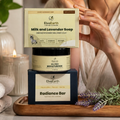 A woman in a beige top applies cream to her face while standing behind a stacked set of ElvaEarth products: a Milk and Lavender Soap box, a Gel Free Moisturizer jar, and a Radiance Bar box, all placed on a wooden tray with candles and lavender sprigs.