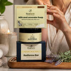 A woman in a beige top applies cream to her face while standing behind a stacked set of ElvaEarth products: a Milk and Lavender Soap box, a Gel Free Moisturizer jar, and a Radiance Bar box, all placed on a wooden tray with candles and lavender sprigs.