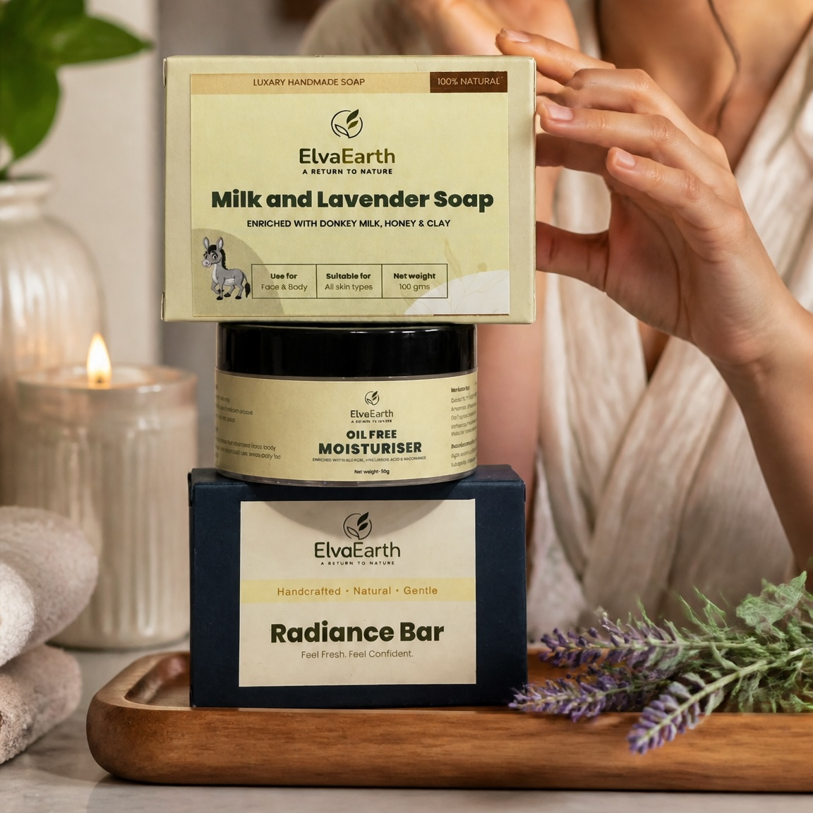 A woman in a beige top applies cream to her face while standing behind a stacked set of ElvaEarth products: a Milk and Lavender Soap box, a Gel Free Moisturizer jar, and a Radiance Bar box, all placed on a wooden tray with candles and lavender sprigs.